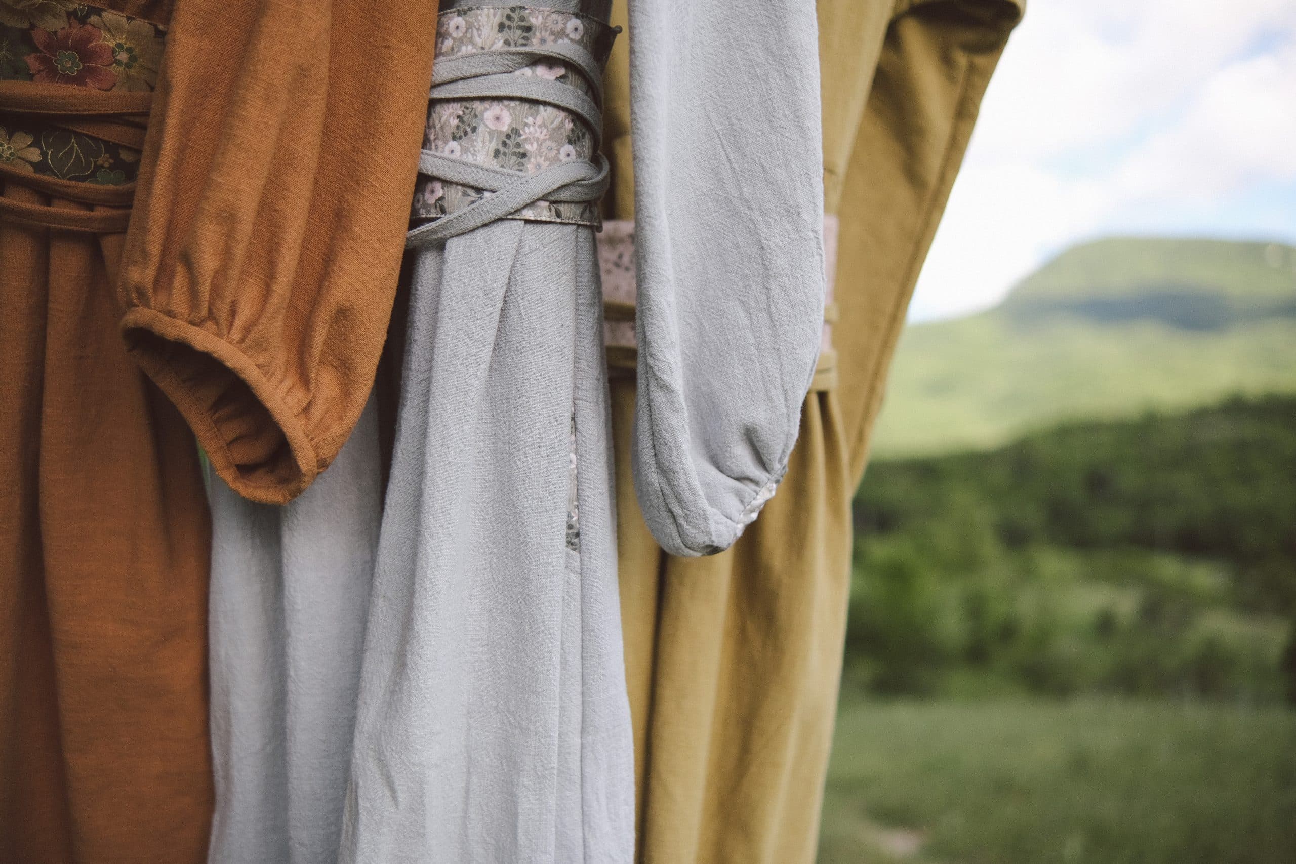 Three dresses — ochre, grey-blue and olive-gold — hanging together, green hills behind