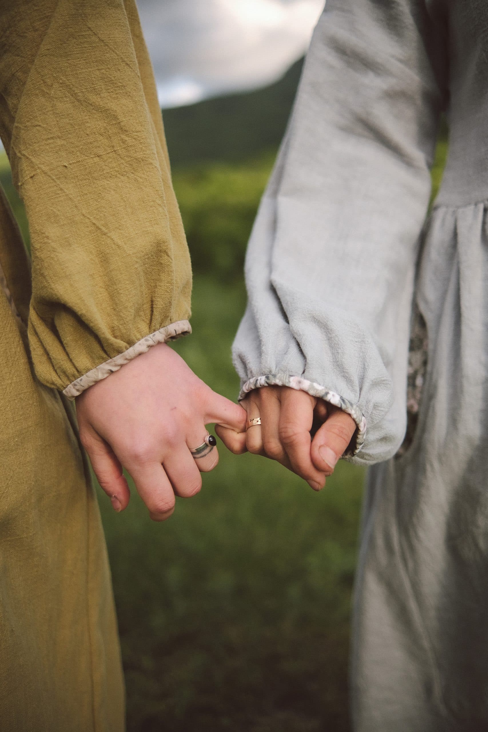Two hands joined by their little fingers — one sleeve olive, one grey-blue, hills behind