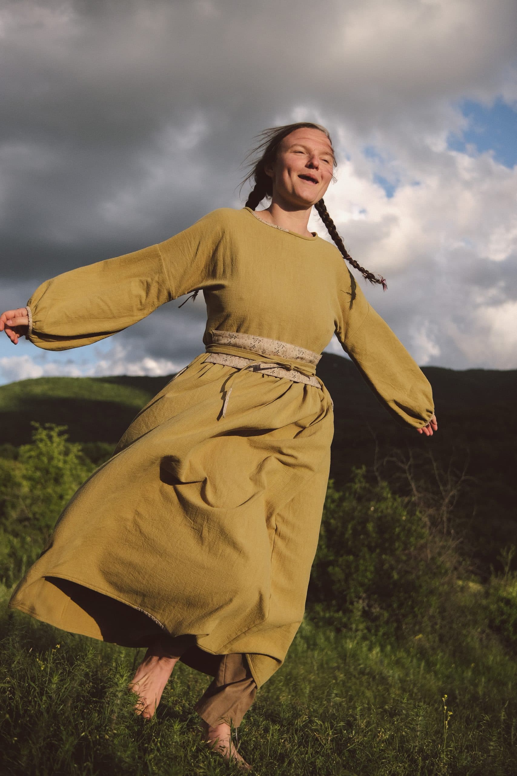 The olive-gold dress mid-turn, skirt swinging against storm-lit hills