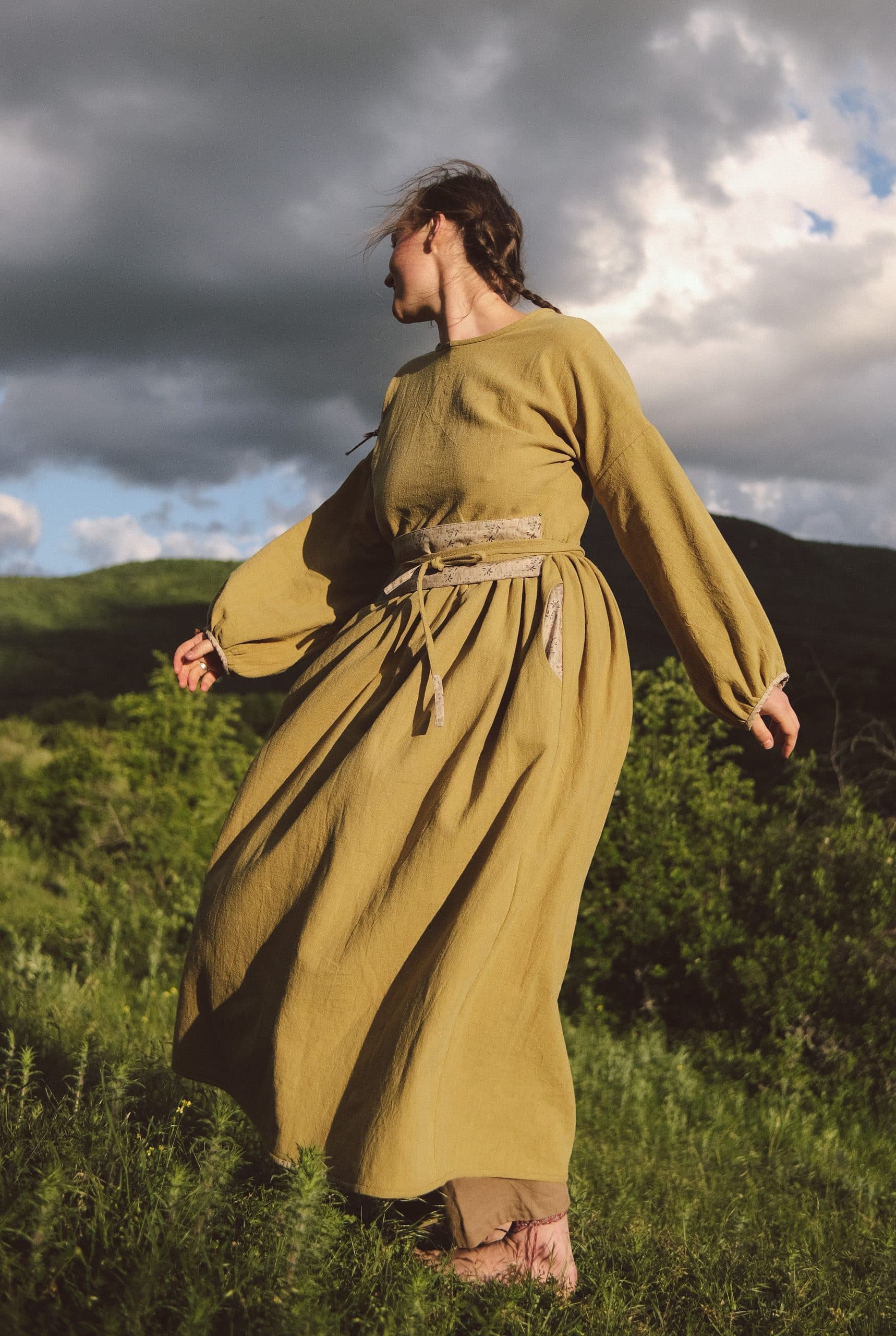 A figure in an olive-gold dress turning in the wind, storm clouds over green hills