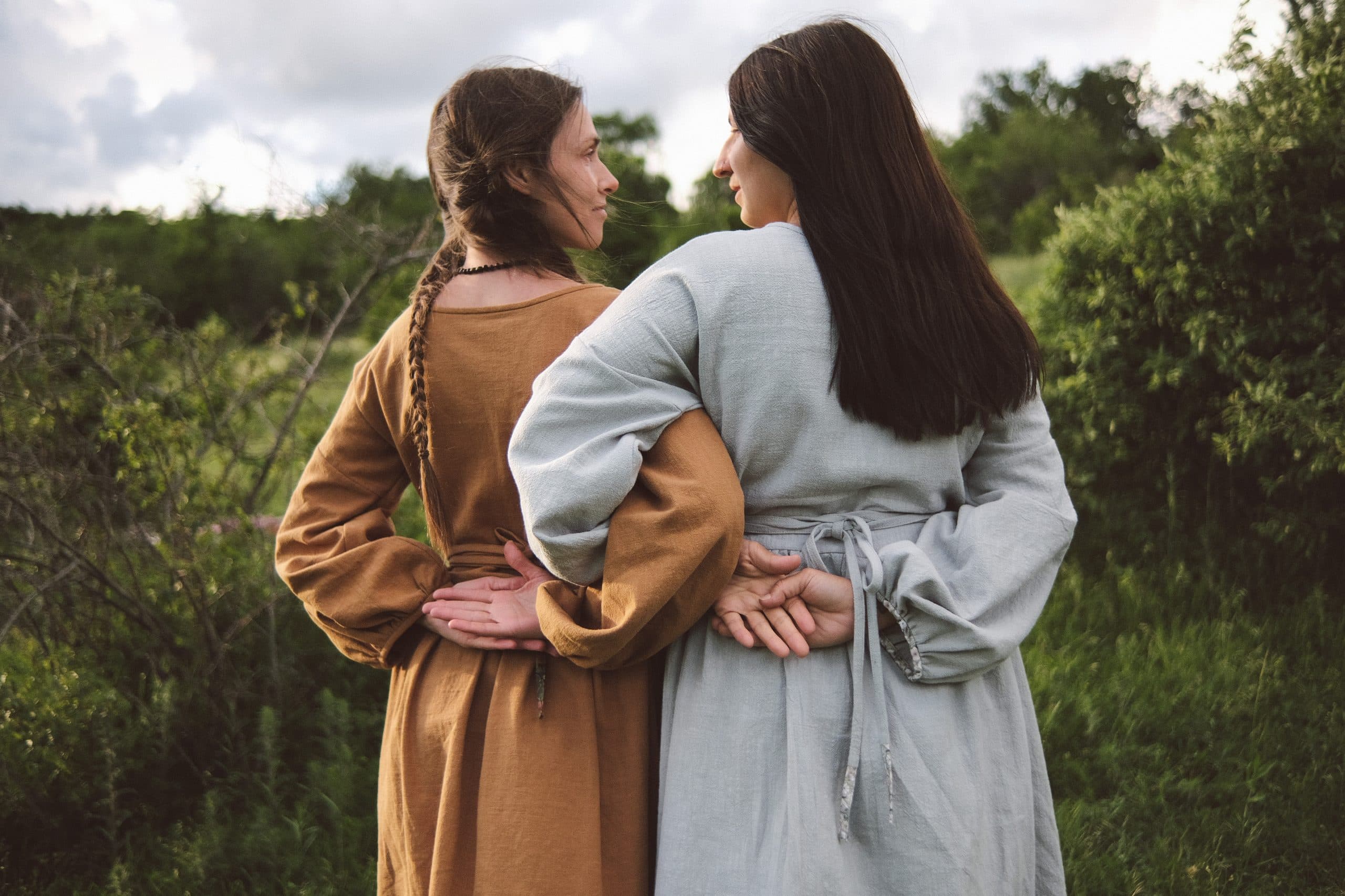 Two dresses seen from behind in a meadow — one ochre, one grey-blue, a sash tied at the back