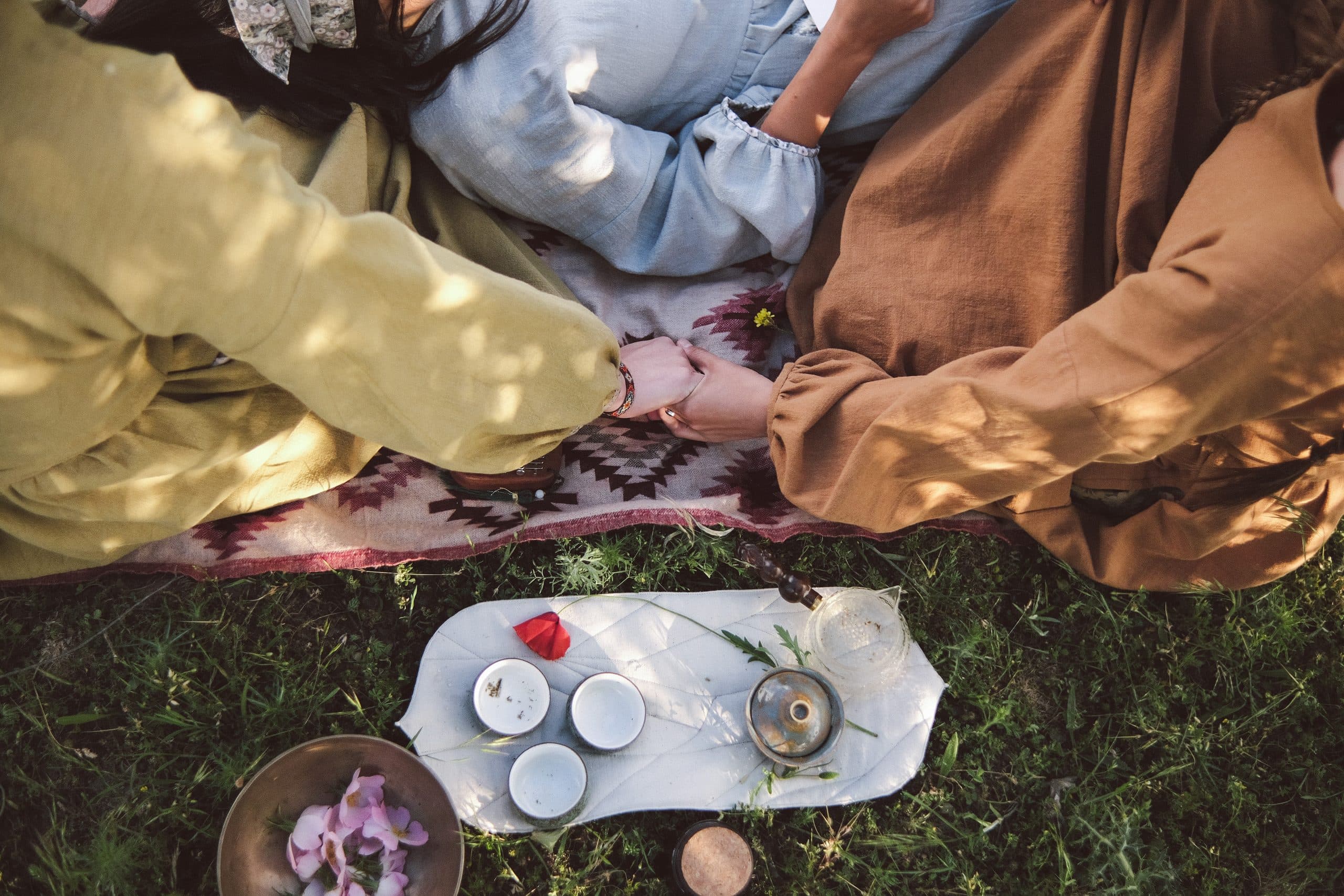 Tea bowls, herbs and joined hands on a patterned rug in the grass, seen from above